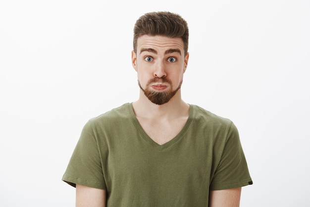 Free photo close-up shot of silly and funny good-looking bearded male in olive t-shirt pouting holding breath looking like balloon raising eyebrows, feeling playful fooling around over white wall