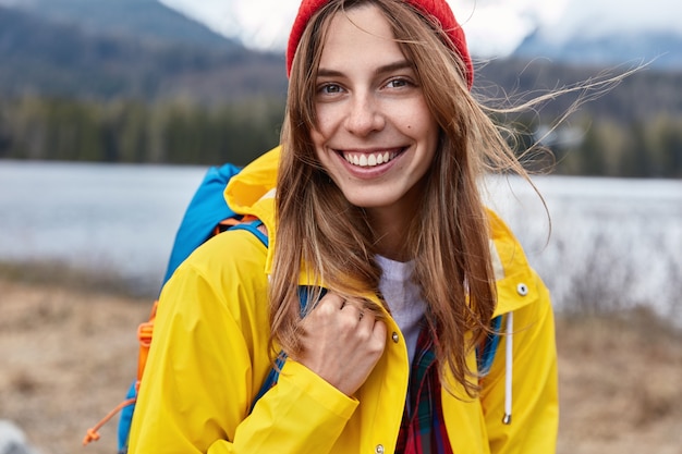 Free Photo close up shot of smiling attractive female tourist in yellow coat, carries rucsack