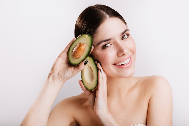 Close-up shot of smiling green-eyed woman without makeup on white wall. Model demonstrates avocado skin benefits.