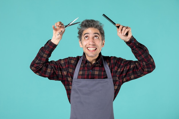 Free photo close up shot of smiling young hairdesser wearing gray apron and cutting his hair on pastel blue color background