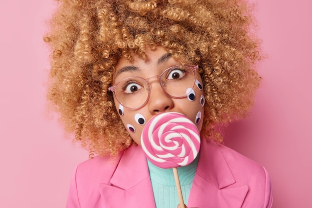 Free photo close up shot of surprised beautiful woman with curly bushy hair holds caramel candy on stick over mouth has widely opened eyes wears transparent eyeglasses formal pink jacket has sweet tooth