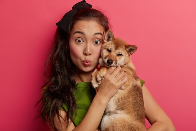 Close up shot of surprised dark haired ethnic woman holds Shiba Inu dog near face, keeps lips rounded, feels closeness with loyal animal