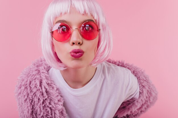 Free Photo close-up shot of surprised young lady in peruke posing with kissing face expression  with pink interior. jocund caucasian girl in periwig enjoying photoshoot