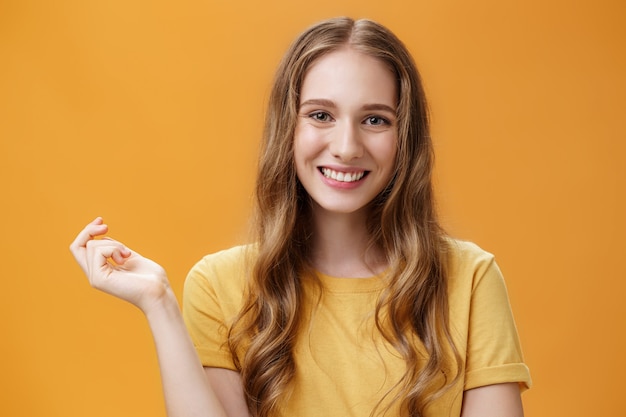 Free photo close-up shot of tender carefree and feminine stylish young woman with natural beautiful wavy hairstyle gesturing with hand over copy space smiling broadly and friendly at camera against orange wall.