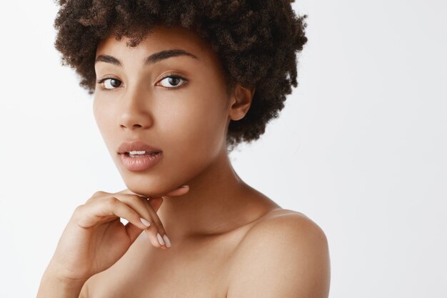 Close-up shot of tender feminine dark-skinned woman with curly hairstyle, touching chin gently with fingers, opening mouth sensually and gazing naked