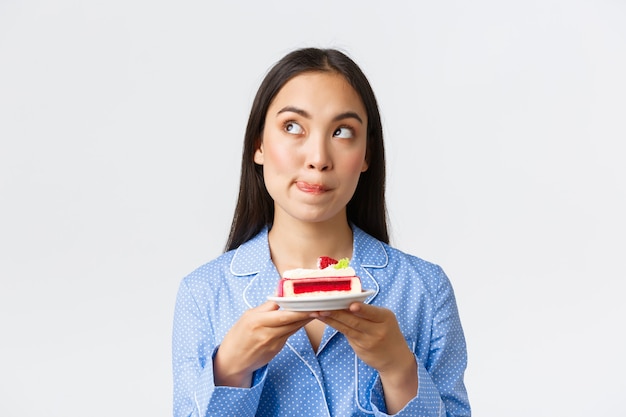 Close-up shot of thoughtful cute asian girl standing at night in pajama with plate of cake, looking upper left corner deciding, cant resist temptation eat delicious dessert, white background