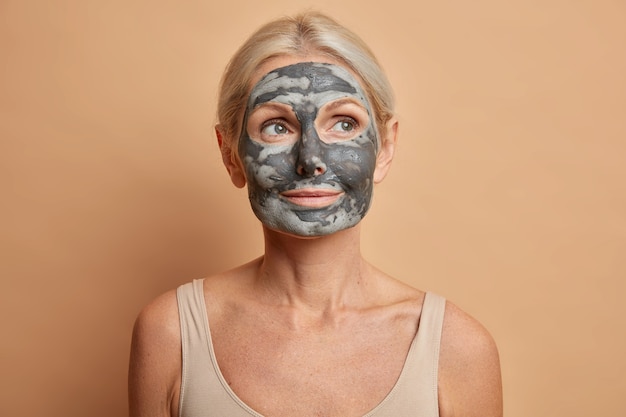 Close up shot of thoughtful European woman applies facial clay mask and looks away cares about complexion poses with bare shoulders isolated on brown wall