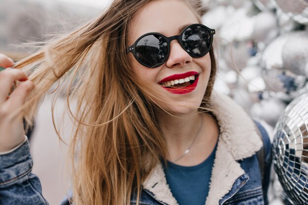 Close-up shot of winsome girl with red lips playing with her hair and laughing on urban street. Photo of pretty caucasian woman in black sunglasses enjoying spring walk.