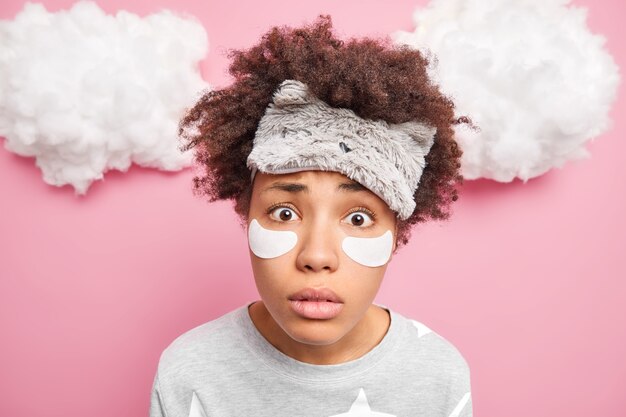 Close up shot of woman stares surprisingly at camera wears sleepmask applies patches under eyes to reduce wrinkles dressed in pajama isolated over pink wall