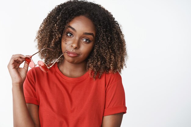 Close-up shot of woman waiting for boyfriend tilting head biting rim of glasses and looking silly and casual at front wearing red bright t-shirt posing doubtful with raised eyebrows over white wall