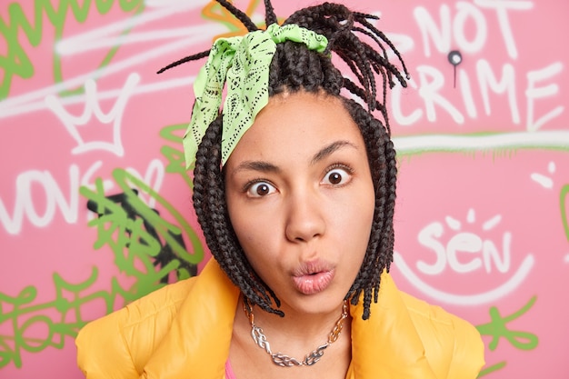 Close up shot of young adult looks with great wonder and surprisement in camera reacts on amazing news has braided hairstyle dressed in vest spends free time at street poses against graffiti wall