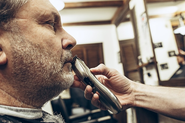 Close-up side profile view portrait of handsome senior bearded caucasian man getting beard grooming in modern barbershop.