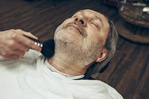 Close-up side top view handsome senior bearded caucasian man getting beard grooming in modern barbershop.