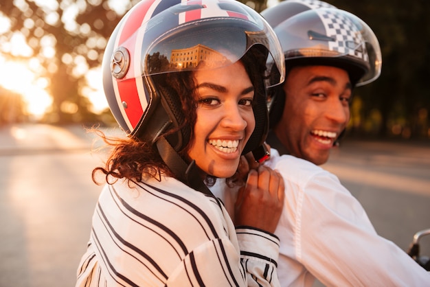 Close up side view image of happy african couple rides on modern motorbike outdoors and looking at the camera