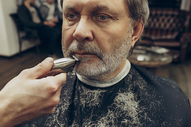 Free photo close-up side view portrait of handsome senior bearded caucasian man getting beard grooming in modern barbershop.