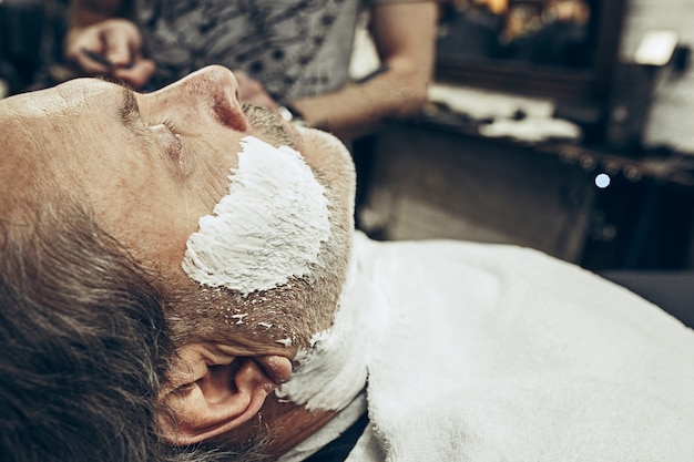 Close-up side view portrait of handsome senior bearded caucasian man getting beard grooming in modern barbershop.