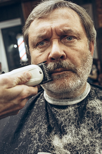 Free photo close-up side view portrait of handsome senior bearded caucasian man getting beard grooming in modern barbershop.