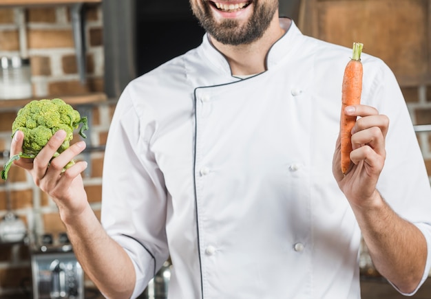 Free photo close-up of smiling chef holding carrot and broccoli in his hand