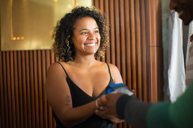 Close-up of smiling female shop assistant at clothing store. Young woman holding credit card reader waiting for payment done looking at shoppers. Clothes business, retail trading and shopping concept