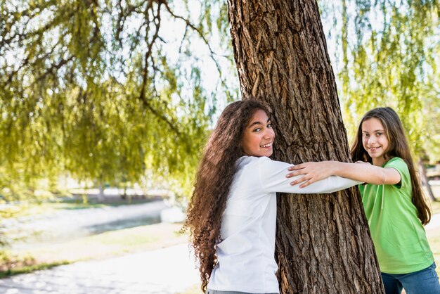 Close-up of smiling girls holding each other's hand hugging big tree in the park