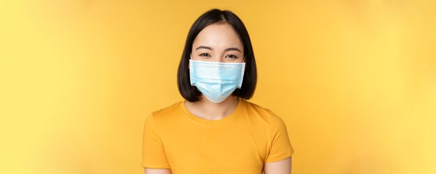 Close up of smiling happy asian woman wearing medical face mask from covid standing in yellow tshirt