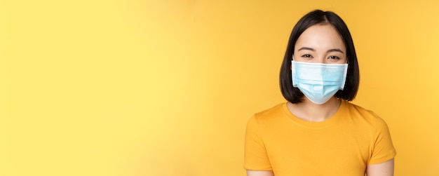 Close up of smiling happy asian woman wearing medical face mask from covid19 standing in yellow tshirt over studio background