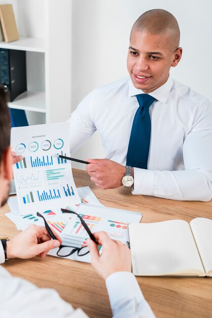 Close-up of a smiling young businessman showing graph with pencil to his business partner at workplace