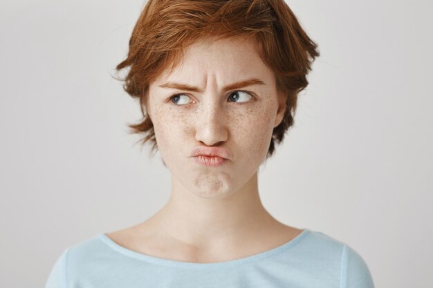 Close-up of sulking and frowning redhead girl posing against the white wall