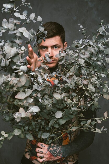 Close-up of a tattooed young man holding dry plant leaves in hand standing against gray background