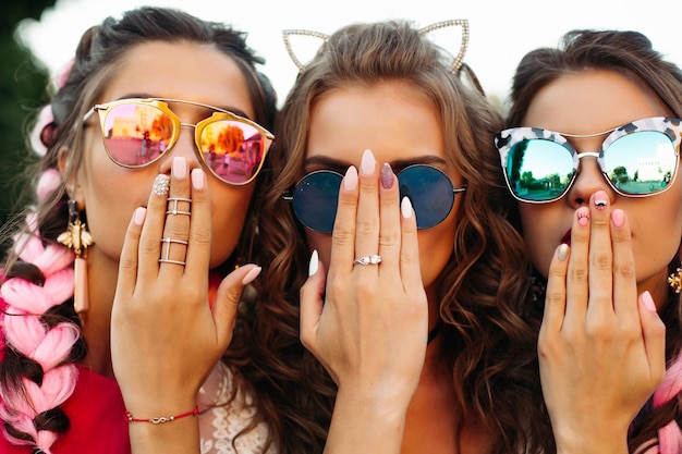 Free photo close up of three young girls wearing in creative sunglasses with designed manicure, two lady covers mouth and one hiding face by hands. happy friends posing and having fun together.