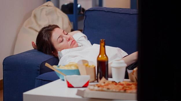 Close up of tired adult laying on couch with TV remote in hand at home. Woman falling asleep on sofa in living room while watching television after eating takeaway fast food meal