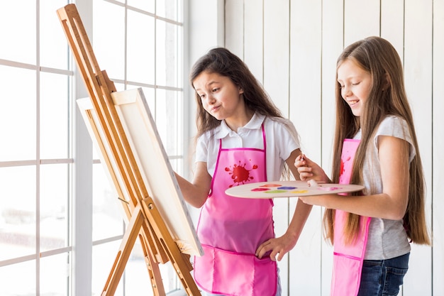 Free photo close-up of two girls standing near the window painting on the easel with paint brush