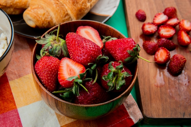 Close-up view of bowl of strawberries with crescent roll in plate on cloth and cut strawberries on cutting board