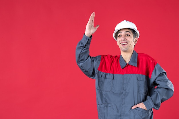 Close up view of confident young architect in uniform with hard hat and pointing up on isolated red wall