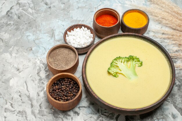 Close up view of creamy broccoli soup in a brown bowl and different spices on gray table