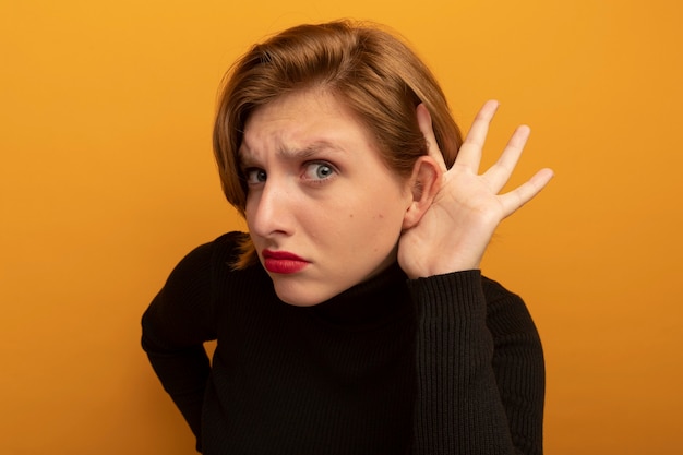 Free photo close-up view of curious young blonde girl  keeping hand on waist putting another hand behind ear doing i can't hear you gesture isolated on orange wall