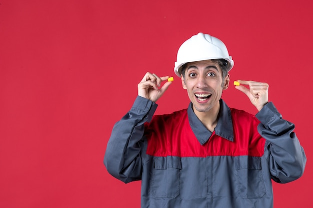 Free Photo close up view of curious young builder in uniform with hard hat and holding earplugs on isolated red wall