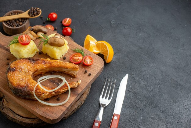 Close up view of delicious fried fish and mushrooms tomatoes greens on cutting board cutlery set pepper on black surface