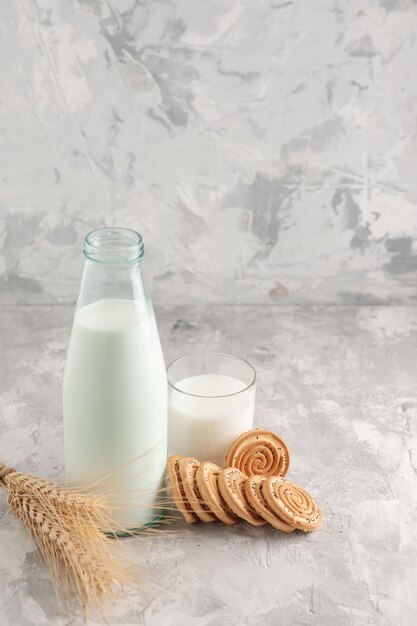 Close up view of glass bottle and cup filled with milk and cookies spike on stained white surface