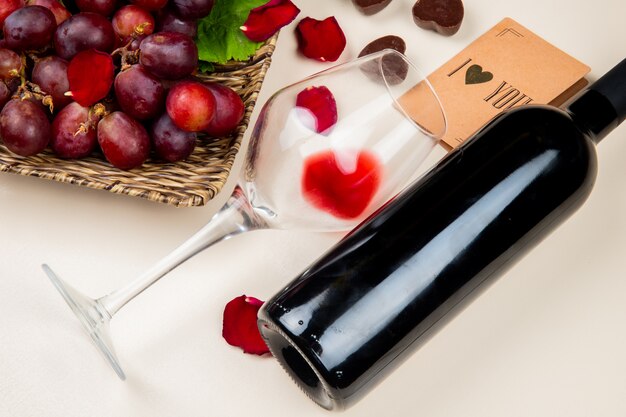 close-up view of glass and bottle of red wine and grape with i love you card on white table decorated with flower petals