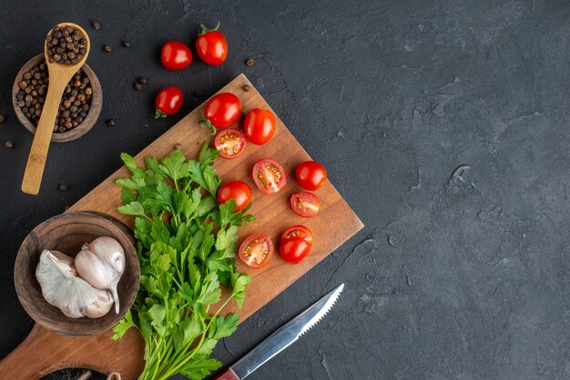 Close up view of green bundle fresh whole cut tomatoes garlics on wooden cutting board knife pepper on black distressed surface