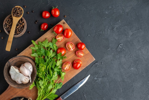 Close up view of green bundle fresh whole cut tomatoes garlics on wooden cutting board knife pepper on black distressed surface