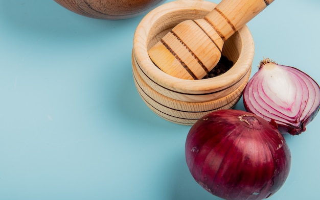 Close-up view of half cut and whole red onions and black pepper seeds in garlic crusher on blue background