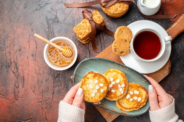 Free photo close up view of hand taking tray with fresh pancakes a cup of black tea on a wooden cutting board honey stacked cookies milk on a dark surface