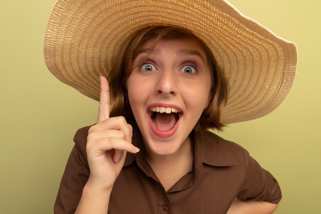 Close-up view of impressed young blonde girl wearing beach hat pointing up 