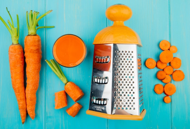 Close-up view of metal grater with carrot juice and whole cut sliced carrots on blue background