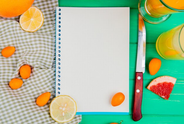 Close-up view of note pad with orange lemon kumquat grapefruit knife and juices around on green background with copy space