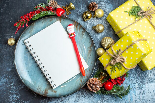 Close up view of notebook with pen and gift boxes on a blue plate decorative accessories conifer cone next to yellow gift boxes on dark background