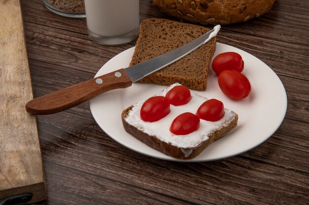 Close-up view of plate of rye bread slices smeared with cottage cheese and tomatoes and knife on wooden background