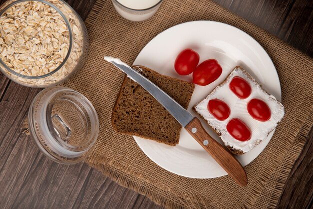 Close-up view of plate with rye bread slices smeared with cottage cheese and tomatoes and knife with oat-flakes on wooden background